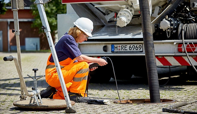 Eine Frau in Arbeitschutzkleidung hockt an einem Schacht und prüft eine Abscheideranlage; im Hintergrund steht ein Saugwagen