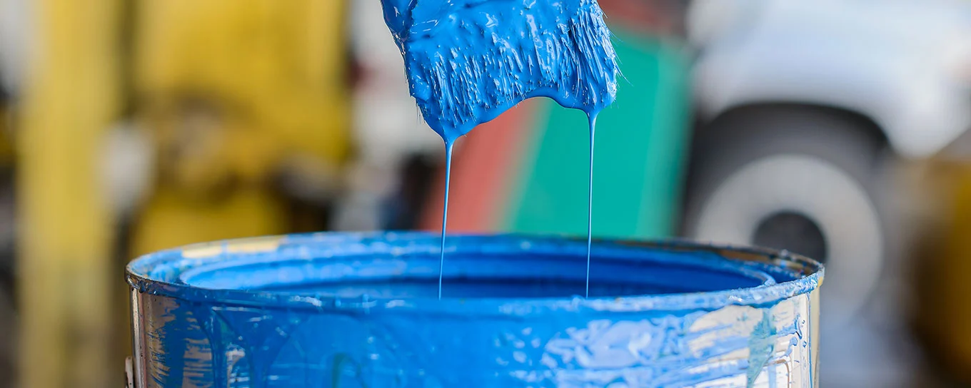 Ein Pinsel wird aus einer Farbdose mit blauer Farbe gezogen und es tropft Farbe vom Pinsel in die Dose.